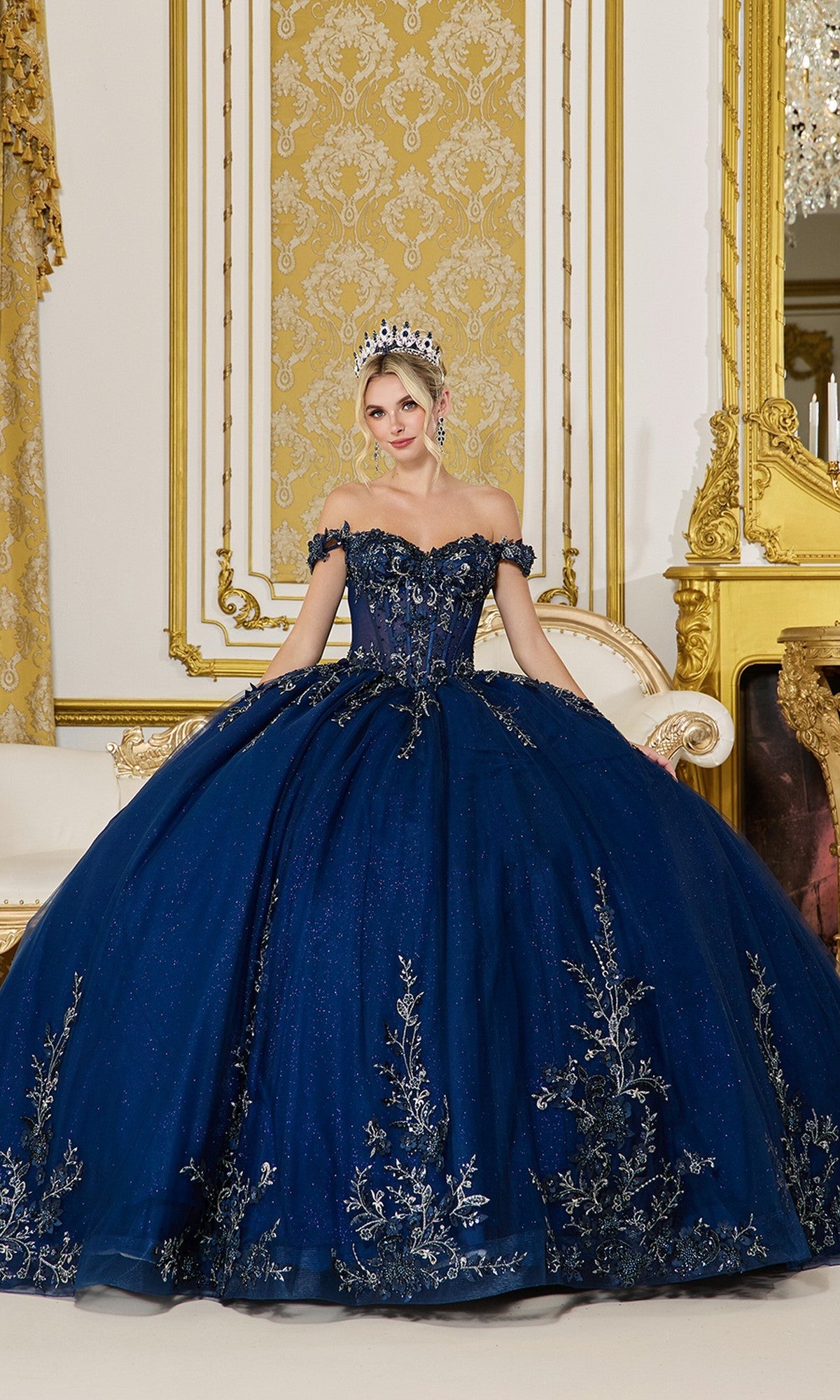 A woman poses in an ornate gold room wearing the Quinceanera Dress 5102 By Dancing Queen in navy, featuring silver lace embroidery and a tiara.